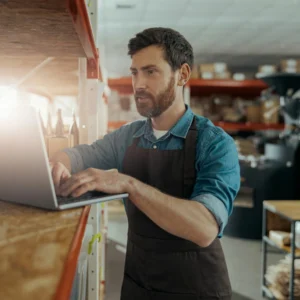 Bearded man in an apron working on a laptop in a warehouse or stockroom.