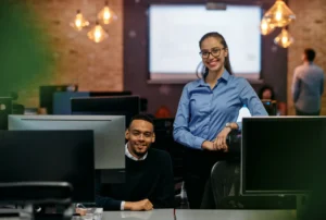 Two colleagues in a modern office smiling confidently in front of computer screens.
