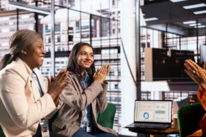 Businesswomen smiling and clapping in front of a laptop showing analytics during a meeting.