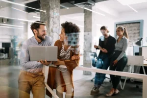 Two coworkers smiling and chatting in a casual modern office setting.