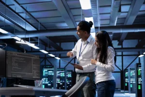 Two IT specialists reviewing code and data on screens in a high-security server room.