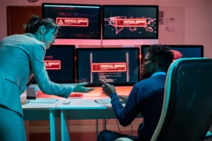 Two cybersecurity professionals reacting to critical error messages on multiple monitors.