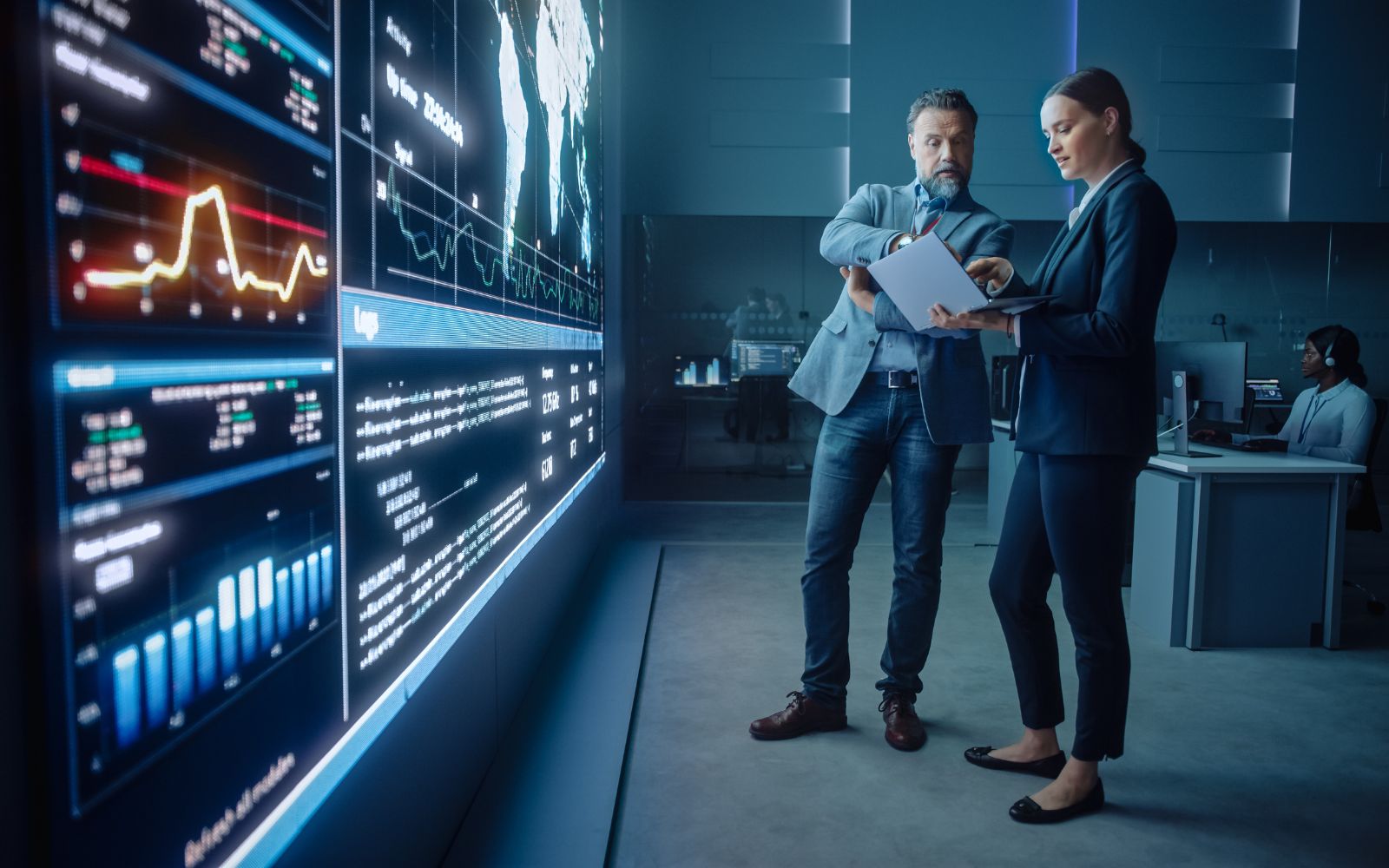 Image of two professionals analyzing data on a large screen displaying charts and maps in a modern office, focusing on data analysis.