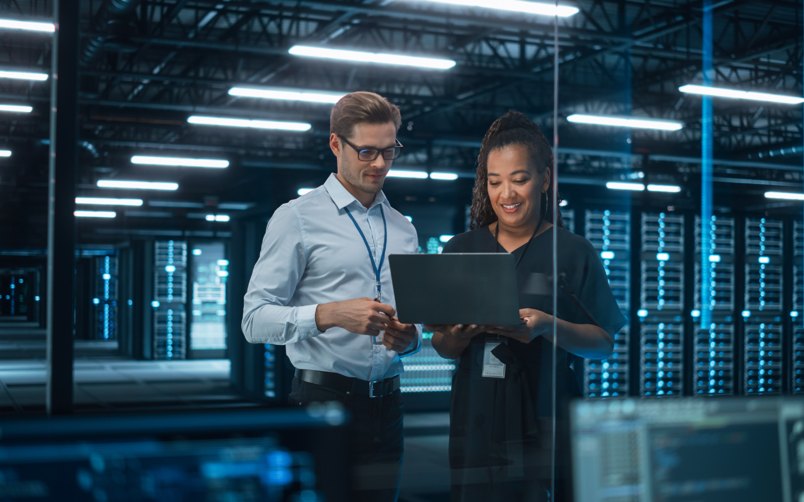 Two professionals collaborating on a laptop in a data center with server racks, discussing IT infrastructure or cybersecurity solutions.